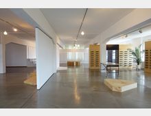 Minimalistisch eingerichteter Innenraum in einem modernen Ausstellungsraum mit weißen Wänden, glattem Betonboden und Holzelementen wie Regalen und Podesten. Im Hintergrund hängen dünne Gardinen vor großen Fenstern, und an der Seite steht eine grüne Zimmerpflanze in einem weißen Topf.