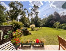 Blick von einer gefliesten Terrasse in einen großen Garten mit weitläufiger Rasenfläche, Blumen- und Pflanzkübeln im Vordergrund sowie einem Holzzaun links. Im Hintergrund stehen hohe Bäume und dichtes Buschwerk unter einem teils bewölkten Himmel.