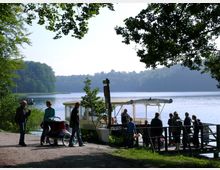 Menschen stehen an einem Seeufer nahe einer Anlegestelle mit einem Boot, das teilweise durch Bäume im Vordergrund eingerahmt ist. Im Hintergrund sind ein bewaldetes Ufer und ruhiges Wasser zu sehen.