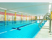 Hallenbad mit einem großen Schwimmbecken, hellblauen Fliesen und gelben Säulen. An den Wänden befinden sich rote geometrische Muster, und die Decke ist mit weißen Paneelen verkleidet.