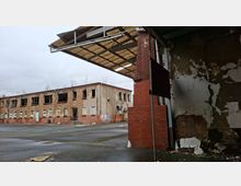Verlassener Industriehof mit einem zweigeschossigen Backsteingebäude, dessen Fenster größtenteils zerstört oder vernagelt sind. Im Vordergrund sind eine bröckelnde Wand, ein roter Ziegelpfeiler und ein teilweise erhaltenes Vordach über einer großen Asphaltfläche zu sehen.