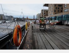 Holzdeck eines Schiffs mit Tischen und Stühlen, im Hintergrund moderne Gebäude aus Backstein und Glas sowie die Oberbaumbrücke mit ihren charakteristischen Türmen. Rettungsringe sind an der Reling befestigt, das Wasser der Spree ist sichtbar.