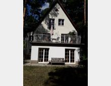 Zweistöckiges Einfamilienhaus mit Satteldach, weißer Fassade und großen Fenstern. Im Erdgeschoss befinden sich Doppeltüren aus Glas, während das obere Stockwerk einen Balkon mit Geländer und eine Sonnenschirmhalterung aufweist. Das Gebäude steht inmitten eines bewaldeten Gartens.