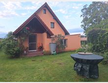 Ein kleines Wohnhaus mit orangefarbener Fassade und einem steilen roten Satteldach. Vor der Haustür befinden sich eine überdachte Terrasse mit Sitzmöbeln sowie Rosenranken an der rechten Wand. Der Garten ist gepflegt, mit einer großen Rasenfläche und dichtem Bewuchs im Hintergrund.