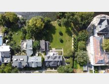 Luftaufnahme einer Wohnsiedlung mit mehreren Gebäuden, die von Gärten und Bäumen umgeben sind. Im Zentrum befindet sich eine große, gepflegte Rasenfläche mit einzelnen Bäumen, und im Hintergrund fließt ein Fluss.