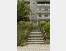 Außenansicht eines Mehrfamilienhauses mit grauer Fassade und Balkonen, davor eine Treppe mit roten Metallgeländern, die von einem Gehweg hinauf zur Eingangstür führt, umgeben von grüner Vegetation.