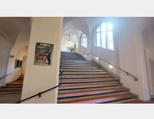 Ein historisch wirkendes Treppenhaus mit breiten Treppenstufen, flankiert von schwarzen Geländern. Rechts befinden sich hohe Fenster mit Rundbögen, die den Raum mit natürlichem Licht durchfluten, während links ein Plakat an einem Pfeiler hängt.