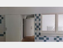 Ein Innenraum mit weißen Wänden und blau-weiß gefliesten Bereichen, einer offenen Tür, die zu einem anderen Raum führt, und einem Heizkörper unter einem Fenster mit breitem Rahmen. Der Boden besteht aus Holzplanken.