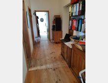 Flur in einem Wohnhaus mit hellem Holzboden und hölzernen Türrahmen. Rechts im Bild befinden sich ein Schrank, Bücherregale mit Aktenordnern sowie ein Laptop; am Ende des Flurs ist ein Dartboard an der Wand zu sehen.