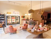 Ein gemütlicher Laden mit hellem Interieur, der eine Mischung aus Café und Boutique bietet. Es gibt eine Kühlvitrine mit Getränken, Regale mit Weinflaschen, eine kleine Sitzecke mit orangefarbenen Sesseln, dekorierte Tische mit Geschenkartikeln und eine beigen Raumteilerwand mit runden Öffnungen im Hintergrund.