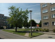 Wohnsiedlung mit mehrstöckigen Apartmenthäusern aus Beton, umgeben von Grünflächen mit Bäumen, Gehwegen und einer Straßenlaterne. Im Hintergrund sind weitere Gebäude mit roten Dächern zu sehen.