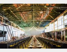 Industriehalle mit hohen Decken, dekoriert mit Lichterketten und langen, festlich gedeckten Tafeln in der Mitte. Große Fenster und eine Holzgalerie an den Seiten sorgen für eine helle und offene Atmosphäre.