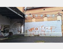 Überdachter Durchgang an einem alten Backsteingebäude mit zugemauerten Rundbogenfenstern und einem unteren Wandbereich aus weißen Fliesen. Die Wände sind teilweise abblätternd und mit Graffiti versehen; der Boden ist asphaltierter Hofbereich.