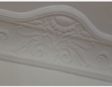 Detail einer weißen Stuckverzierung mit geschwungenen floralen Mustern und einer Blütenform in der Mitte. Die Verzierung befindet sich an einer Wand oder Decke mit leicht gewellter Umrahmung.