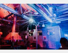 Industrieller Veranstaltungsraum mit sichtbaren Stahlträgern, Lüftungsrohren und einer Discokugel, die Lichtpunkte an die Wände wirft. Der Raum ist in bunten, überwiegend roten und blauen Lichtern beleuchtet und mit Ballons sowie Dekoration für eine Feier geschmückt.