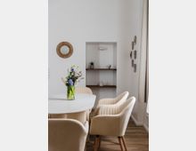 Eine helle, minimalistisch eingerichtete Ecke eines Wohnzimmers mit einem weißen Tisch, umgeben von cremefarbenen Polsterstühlen. Im Hintergrund befinden sich eine Vertiefung mit Holzregalen, auf denen Dekorationsobjekte stehen, sowie eine runde Wandspiegel und kleine Wandbilder.