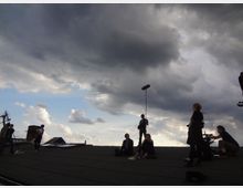 Gruppe von Menschen auf einem Flachdach unter einer bewölkten Himmelsszenerie, mit Film- oder Tonaufnahme-Equipment wie Mikrofonen und Kameras sichtbar. Im Hintergrund sind Wolken in verschiedenen Grautönen mit kleinen blauen Stellen am Himmel zu erkennen.