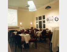 Ein Veranstaltungsraum mit cremefarbenen Wänden, einem großen Fenster und einer Projektionsfläche. Der Raum ist mit runden Tischen und roten Stühlen möbliert, die Tische sind mit weißen Tischdecken und Blumenschmuck dekoriert.