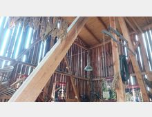 Innenansicht einer rustikalen Scheune mit Holzbalken und vertikal angeordneten Holzwänden, durch die Licht einfällt. Im Raum hängen getrocknete Blätter und landwirtschaftliche Geräte, darunter ein Wagenrad und ein Geschirr, sowie Poster mit Informationen an den Wänden.