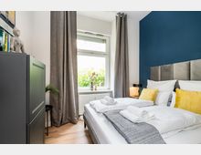 Modernes Schlafzimmer mit Doppelbett, grauem gepolstertem Kopfteil und blauer Akzentwand, weißer Bettwäsche und gelben Kissen. Großes Fenster mit langen grauen Vorhängen über einem Heizkörper sowie ein dunkler Schrank und eine kleine Nachttischlampe.