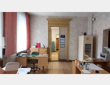 Historisches Büro mit einfacher Möblierung, bestehend aus Holztischen, Bürostühlen und einem Kachelofen an der rechten Wand. Im Hintergrund ein Durchgang mit einer rahmenden Holztür, der in einen weiteren Raum mit Schränken und einer metallenen Aktenschrank führt.