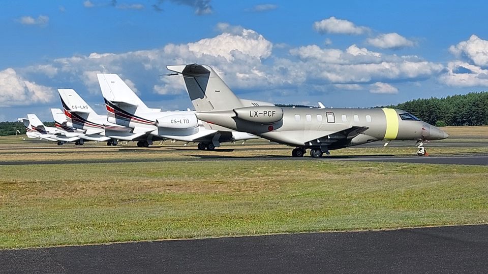 Abstellfläche auf einem Flughafen mit mehreren geparkten Geschäftsflugzeugen in einer Reihe auf dem Rollfeld, davor Grasstreifen und Asphalt. Im Hintergrund sind eine Baumlinie und ein blauer Himmel mit Wolken zu sehen.