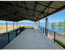 Überdachte Aussichtsplattform aus Holz mit Metallgeländern, die auf Stelzen über einer Schilflandschaft und einem See mit klarem, blauem Wasser errichtet ist; im Hintergrund sind grüne Bäume und der Horizont sichtbar.