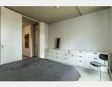 Minimalistisch eingerichtetes Schlafzimmer mit Betondecke, weiß gestrichenen Wänden und einem großen Bett mit schwarz-weiß gemusterter Decke. Im Raum befinden sich eine lange, weiße Kommode mit Schubladen, ein schwarzer Stuhl, ein an die Wand gelehnter Spiegel sowie geöffnete Türen, die zu einem angrenzenden Flur führen.