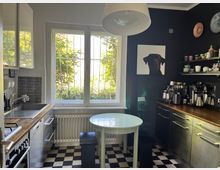 Kleine Küche mit schwarz-weißem Schachbrettmuster-Boden, weißen Wänden, einer großen Fensterfront mit Gitter und einem weißen Heizkörper darunter. Links befinden sich eine Spüle, ein Herd und Schränke, rechts eine dunkle Arbeitsplatte mit Kaffeegeräten und offenen Regalen. An der Wand hängt ein Bild eines schwarzen Hundes.