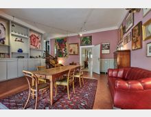 Wohnzimmer mit rosa Wänden, dekoriert mit bunten Gemälden und Bildern. Der Raum enthält einen Holzesstisch mit Stühlen, ein rotes Ledersofa, einen Teppich mit orientalischem Muster sowie Regale mit Büchern und Dekorationsgegenständen. Im Hintergrund sind eine Treppe und geöffnete Türen zu sehen.