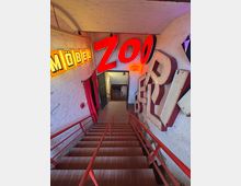 Treppenabgang in einem Retro-Industrie-Interieur mit verklinkerten Wänden, roten Geländern und großformatigen, leuchtenden Reklamebuchstaben an den Wänden, darunter Schriftzüge wie „Möbel“ und „Zoo“. Am unteren Ende der Treppe ist ein kleiner Durchgang sichtbar, teilweise mit Vorhängen bedeckt.