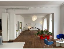 Großer, offener Arbeitsbereich mit weißen Wänden und Holzboden. Im Raum befinden sich ein langer Holztisch mit Stühlen, eine rote Pflanzentruhe mit grünen Blättern, verschiedene Designstühle und eine weiße Stehlampe, beleuchtet durch natürliche Fensterbeleuchtung und eine zentrale Papierlampe.