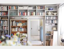 Wohnraum mit einer Wand aus hellgrauen Bücherregalen, die mit Büchern, CDs, DVDs und Dekorationsstücken gefüllt sind. In der Mitte befindet sich ein Türrahmen, der in einen hellen Flur führt; unten links sind verwischte Blumen im Vordergrund sichtbar.