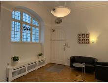 Heller Eingangsbereich mit weiß gestrichenen Wänden, einem großen Fenster mit Buntglasdetails, einer weißen Holztür und einem Holzboden im Fischgrätmuster. Im Raum stehen schwarze Ledersessel, ein kleiner Glastisch und Pflanzen auf einer Heizungskonsole. Ein Bild mit Schmetterlingsmotiven und eine Wandlampe schmücken die Wand.