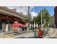Straßenansicht mit einem roten Backsteingebäude, das ein Café im Erdgeschoss beherbergt, mit roten und weißen Sonnenschirmen und Sitzgelegenheiten im Außenbereich. Im Vordergrund stehen Fahrräder an Ständern und ein orangefarbener Mülleimer, während im Hintergrund Bäume und moderne Stadtarchitektur zu sehen sind.