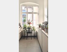 Heller Küchenraum mit einer großen, bogenförmigen Fensterfront, die Tageslicht hereinlässt. Unter dem Fenster stehen zahlreiche grüne Pflanzen in Töpfen, daneben eine schwarze Stuhlgestaltung; helle Schränke und Arbeitsflächen säumen die Seiten.