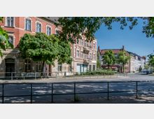 Straßenszene mit historischen Wohnhäusern aus rotem Backstein und dekorativen weißen Fassadenelementen, flankiert von Bäumen. Im Vordergrund ein Gehweg und ein Geländer, im Hintergrund weitere Gebäude und eine Straße mit geparkten Autos.