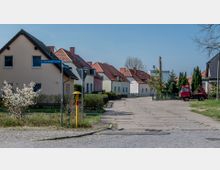 Wohngegend mit Einfamilienhäusern, die pastellfarbene Fassaden und rote Ziegeldächer haben. Im Vordergrund befindet sich eine kleine gepflasterte Seitenstraße mit einem Straßenschild, einem blühenden Strauch und einem roten Transportfahrzeug; im Hintergrund ist ein Industriekamin zu sehen.