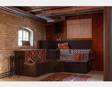 Gemütlicher Raum mit rustikalem Backsteinmauerwerk, einer kleinen Fensteröffnung und sichtbaren Metallrohren an der Decke. Die Sitzecke ist mit schwarzen Möbeln, bunten gemusterten Decken und Kissen gestaltet; auf dem Boden liegt ein farbiger Teppich.