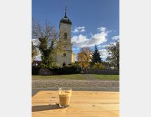 Blick auf eine helle Kirche mit quadratischem Turm und dunkler Kuppel, umgeben von Bäumen und einer niedrigen Steinmauer unter blauem Himmel mit Wolken. Im Vordergrund steht auf einem Holztisch ein Glas Kaffee, dahinter ein gepflasterter Platz.
