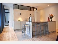 Moderne Küche mit hellem Holzfußboden, einer Kücheninsel mit grauen Barhockern und einer integrierten Arbeitsplatte. Die Rückwand besteht aus beigefarbenem Putz und ist durch schwarze Fensterrahmen mit einem Oberlicht abgesetzt.