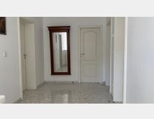 Flur mit weißen Wänden und hellgrauem, marmoriertem Fliesenboden; zentral ein großer, wandmontierter Spiegel mit dunklem Holzrahmen, daneben eine geschlossene, hellbeige Tür mit Zierkontur. Links und rechts führen weitere Türen in angrenzende Räume.