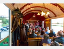 Innenraum eines historischen, hölzernen Wagens mit gewölbter Decke und roten Vorhängen, in dem eine Theateraufführung oder Lesung vor einem sitzenden Publikum stattfindet. Im Hintergrund stehen mehrere Schauspieler in historischer Kleidung auf einer kleinen Bühne, während draußen parkende Autos sowie eine Landschaft mit Wiese und Bäumen sichtbar sind.