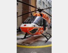 Ein orange-weiß lackierter Rettungshubschrauber steht auf einer markierten Landefläche in einem Innenhof, umgeben von Gebäuden mit Backstein- und Putzfassaden. Die Rotorenblätter des Hubschraubers sind ausgeklappt.