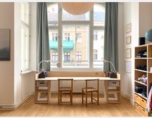 Heller Arbeitsbereich mit langem Holztisch und zwei Stühlen vor großen Sprossenfenstern mit Vorhängen, ausgestattet mit Schreibtischlampen und Stifteboxen. Rechts steht ein offenes Regalsystem mit Fächern, links sind weitere Fenster und ein Wandbild zu sehen.