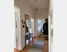 Ein schmaler Flur mit Holzdielenboden und weißen Wänden. An der linken Wand hängt ein goldgerahmter Spiegel über einer kleinen Kommode, daneben sind gerahmte Fotos angebracht. Am Ende des Flurs sind zwei offene Türen sichtbar, und rechts ist ein Kleiderständer mit Kleidung.