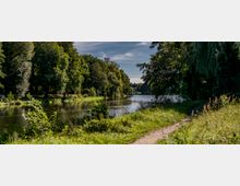 Ein Fluss in einer natürlichen Umgebung, umgeben von dichtem Grün und großen Bäumen, mit einem schmalen, grasbewachsenen Pfad am rechten Ufer. Der Himmel ist teilweise bedeckt, und das Wasser reflektiert die umliegende Vegetation.