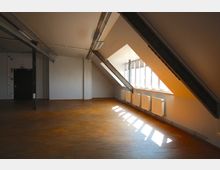 Ein leerer Dachraum mit schrägen Decken, sichtbaren Stahlträgern und einem Holzboden. Rechts befinden sich große Fenster mit Heizkörpern darunter, durch die Tageslicht einfällt; links eine schwarze Tür.