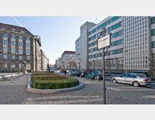 Straßenplatz in einer Stadt mit Gehweg aus Pflastersteinen, einem kleinen rechteckigen Grünbereich mit Hecke und umliegenden Parkbänken. Im Hintergrund historische und moderne Gebäude sowie parkende Autos; ein Schild mit der Aufschrift „Zietenplatz“ ist im Vordergrund sichtbar.