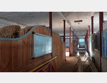 Ein langer Pferdestall mit weißen Deckenbalken und roten Metallstützen. Links sind Holzboxen mit Heuballen und einer Informationstafel, rechts befinden sich geschlossene Metallboxen; am Ende des Gangs ist ein Fenster mit Gitter zu sehen.